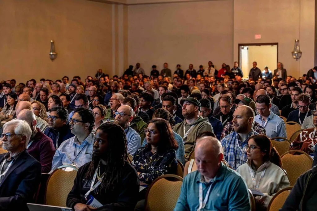 A seated breakout audience listening, with more people standing along the back wall