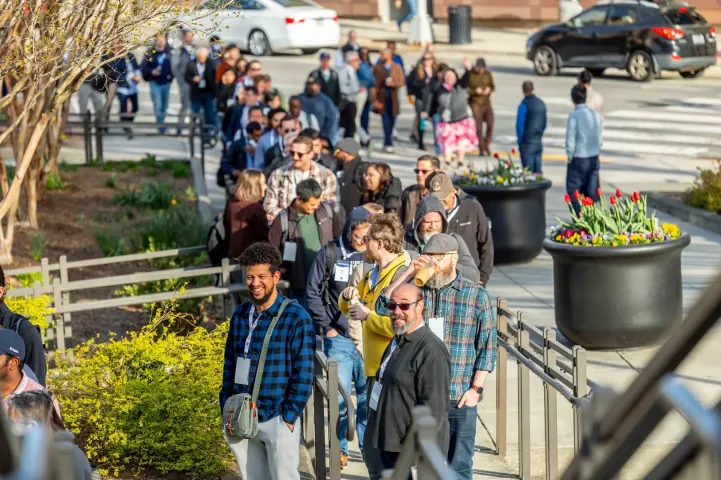 A long line of smiling attendees waiting outside the 2026 venue in spring sunshine; the 2027 venue in Raleigh has room for everyone