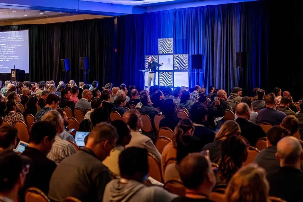 A speaker on stage presenting to a packed room, some attendees following along on laptops