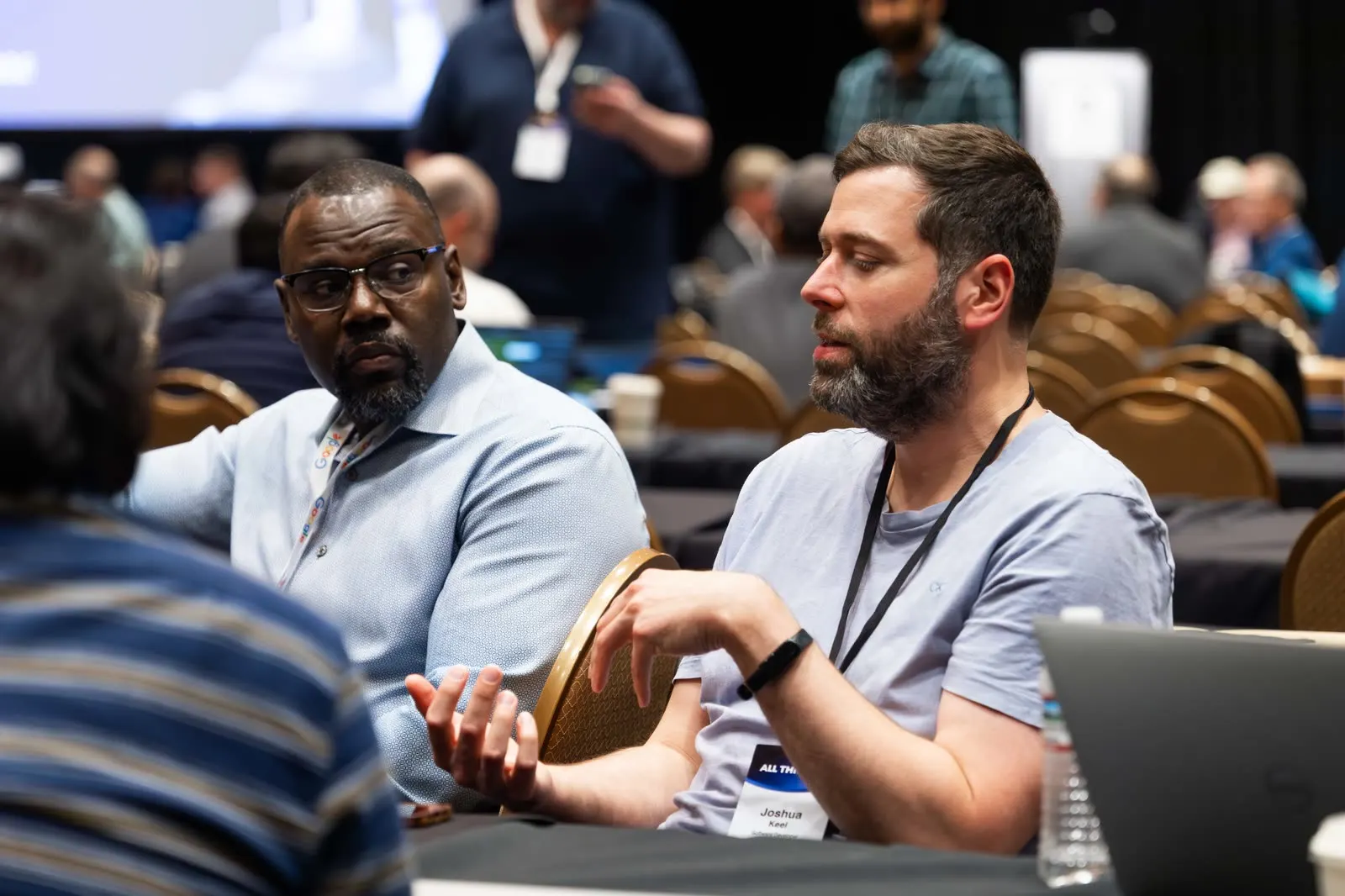 Two people at a workshop table at All Things AI 2026, one explaining something with his hands while the other listens