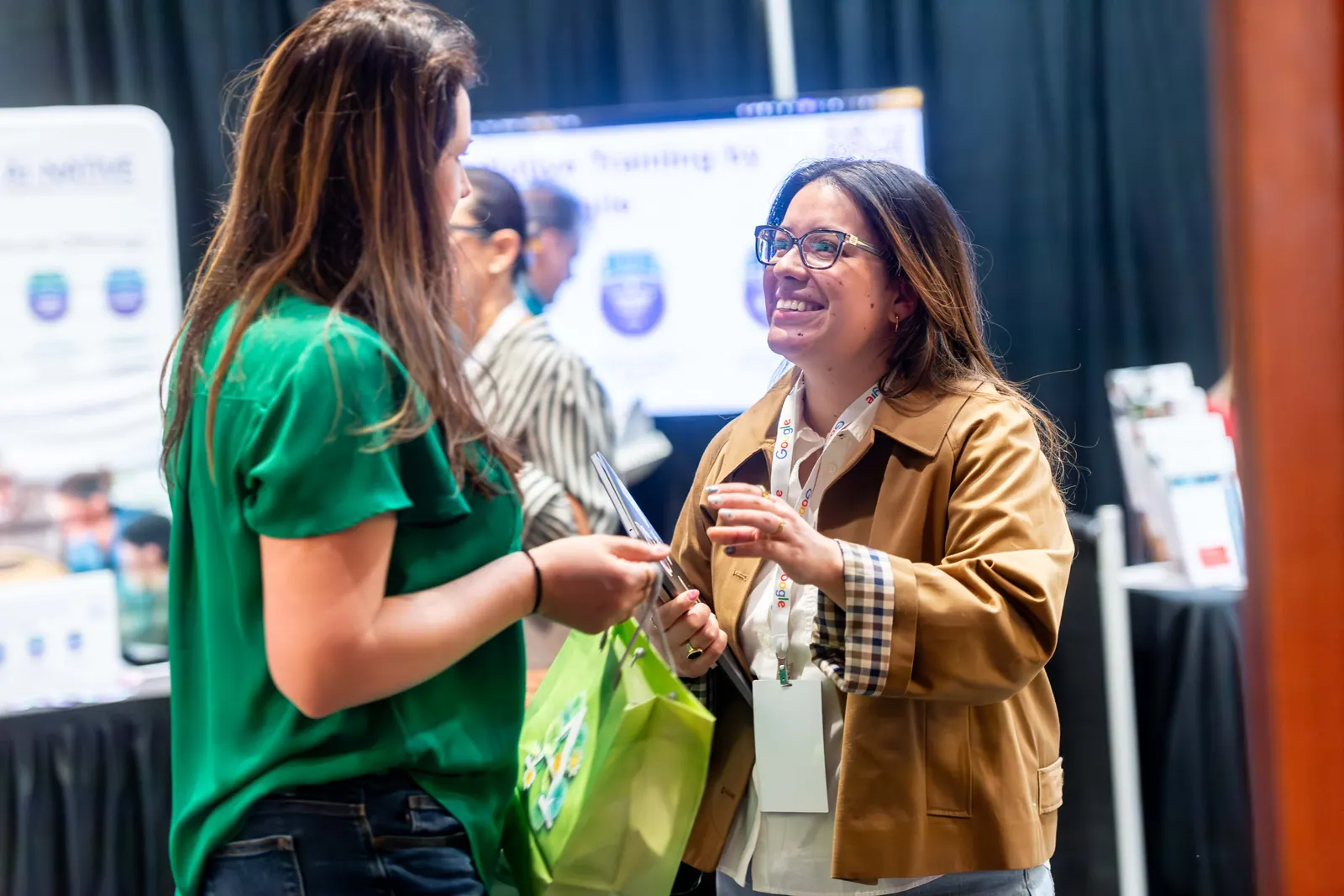 Two people laughing together at a sponsor table at All Things AI 2026, one holding a green swag bag