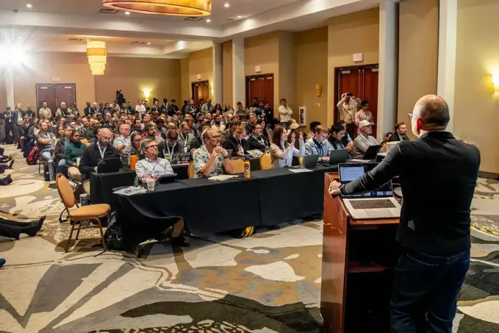 A speaker at the podium in the foreground, a full room listening in front of him