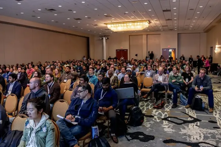 A packed and diverse breakout room of business executives, developers, knowledge workers and AI engineers, every chair taken and more attendees on the floor