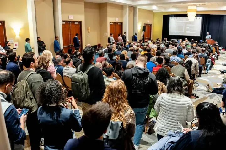 An overflow crowd standing and sitting in the aisles to see a 2026 session; rooms like this are why 2027 moves to a bigger venue