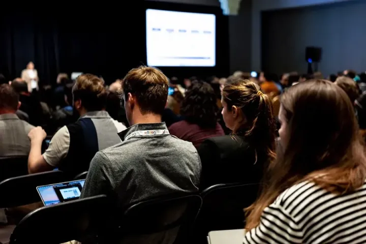 Attendees in a dark session room following the slides, laptops on their knees