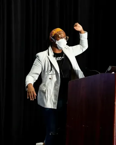 A speaker in a white blazer raising one arm high behind the podium, asking the room to raise their hands