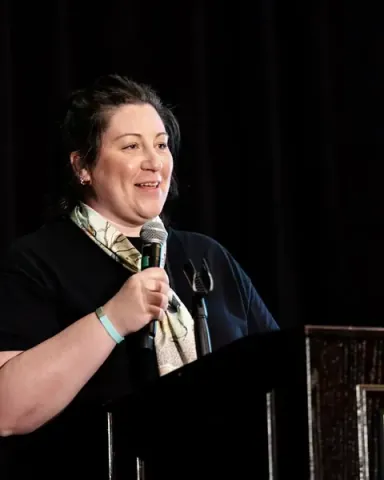 Sarah Chick of NPower North Carolina smiling at the audience from the podium, microphone in hand