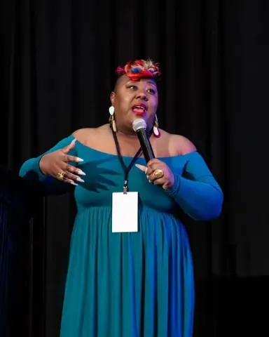 Chisa Pennix-Brown gesturing to the room on stage in a teal dress, microphone in hand