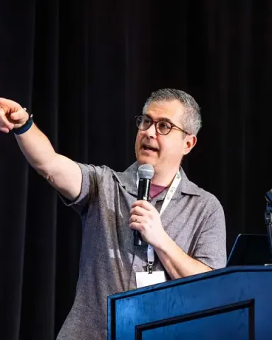 Marc Sirkin pointing out into the audience from the podium, microphone in hand