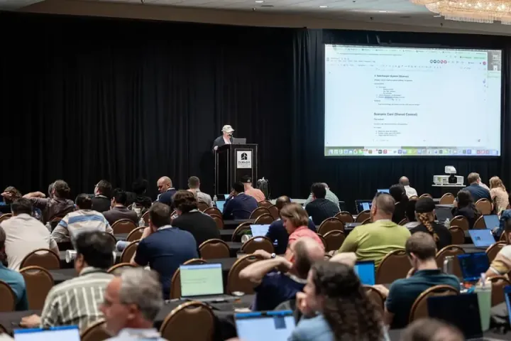 John Willis leading an AI DevSecOps workshop, a large room working along on laptops