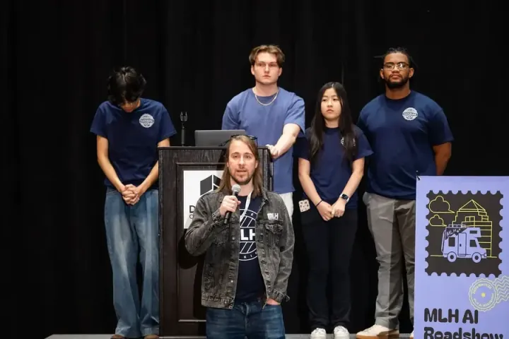 Major League Hacking presenting the MLH AI Roadshow on stage, a speaker at the microphone with the student team behind him