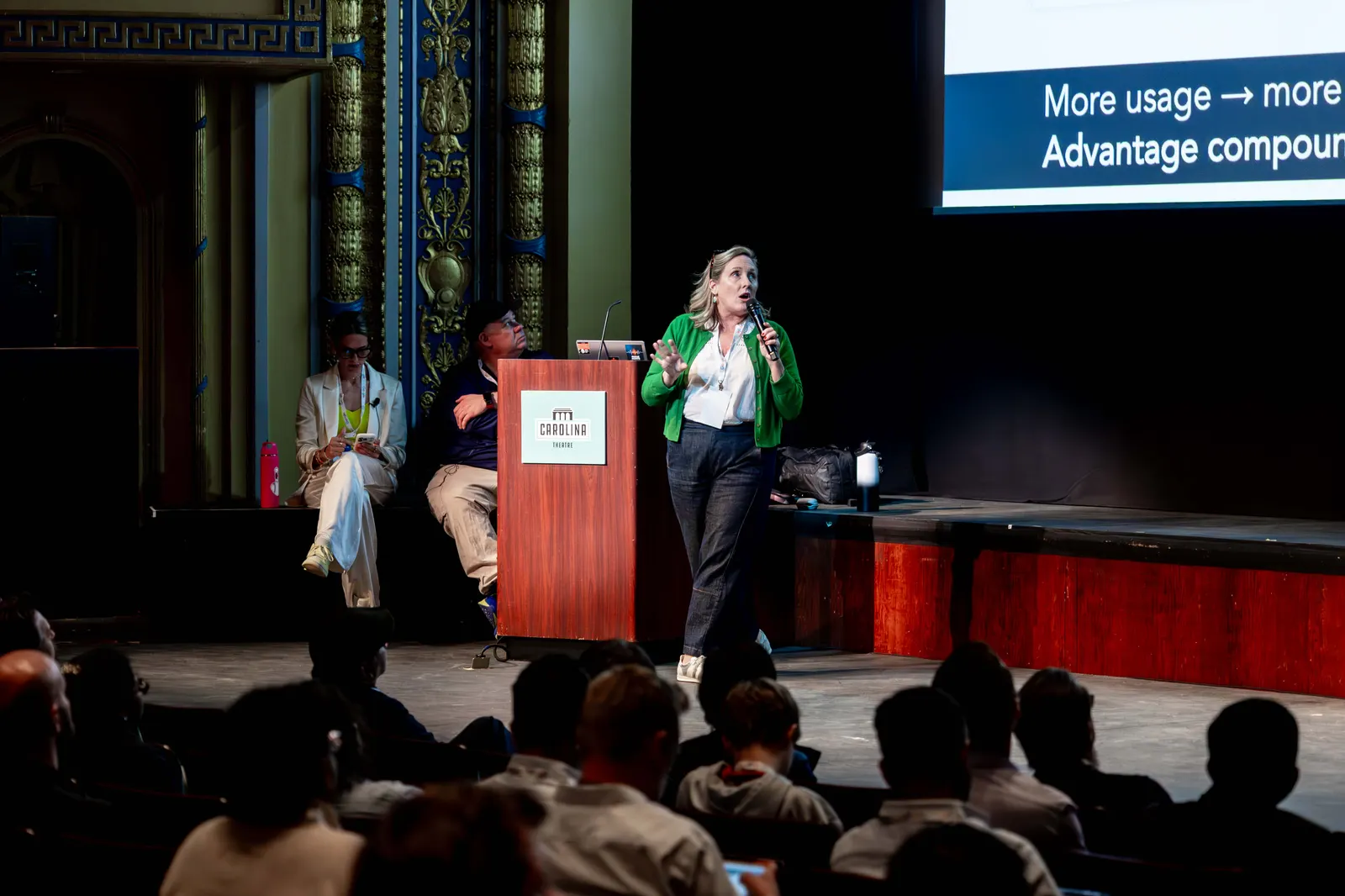 A speaker presenting beside a large screen on the Carolina Theatre stage at All Things AI 2026