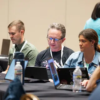 Three people working side by side at a workshop table, laptops open