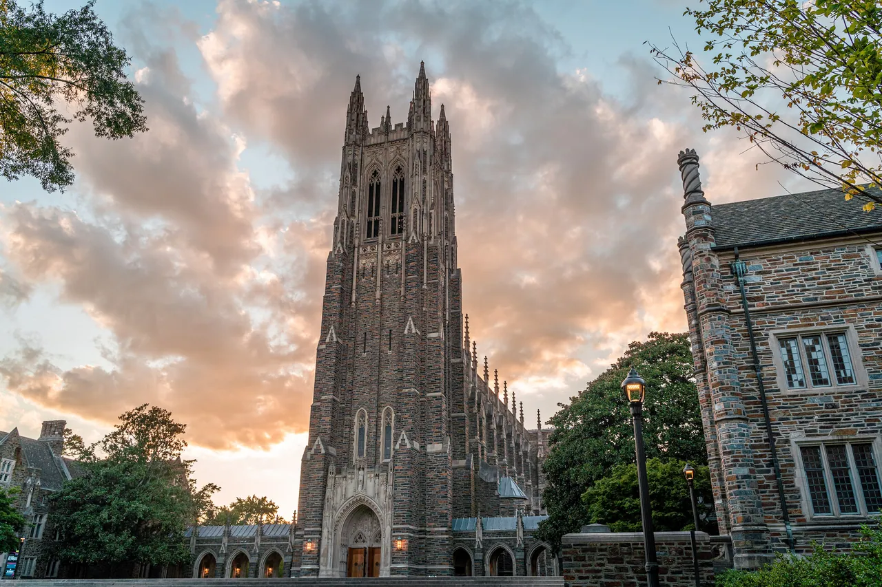 The stone tower of Duke University Chapel rising against an orange and blue sunset sky, framed by trees and campus buildings.