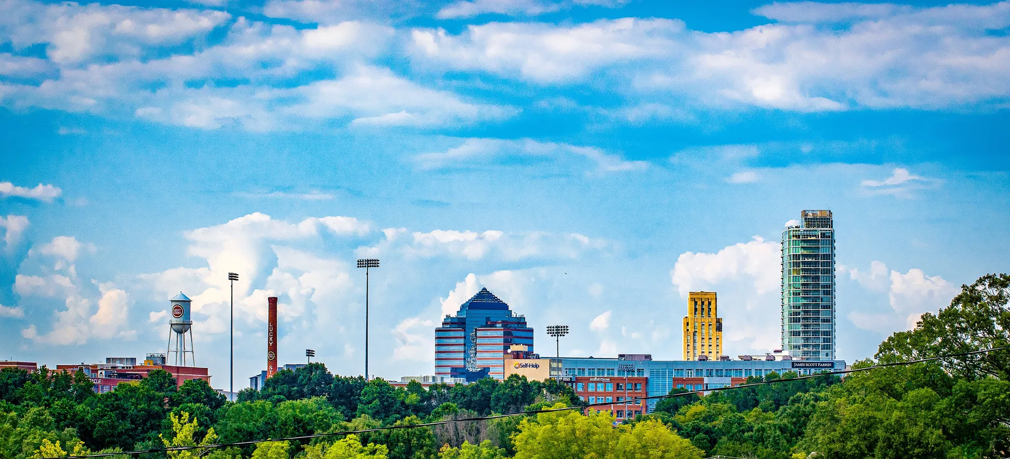 The downtown Durham skyline under a blue sky with scattered clouds: a blue glass office building, a gold Art Deco tower and a tall glass tower rising above green trees, with a brick smokestack at the far left.