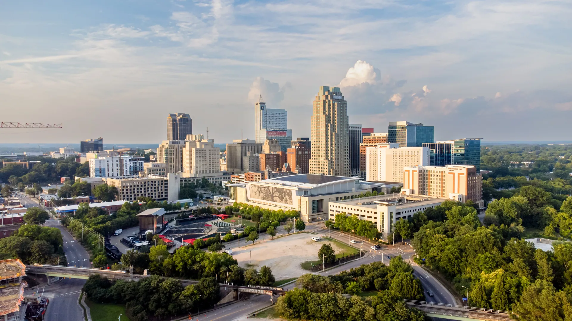 Downtown Raleigh at golden hour, seen from Dorothea Dix Park: office towers and mid-rise buildings above tree-lined streets, with soft clouds behind.
